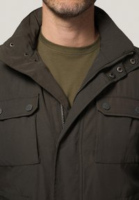 Ciemnobrązowa kurtka z zamkiem błyskawicznym, zatrzaskami i dwoma kieszeniami na piersiach. Nosi się ją na oliwkowej koszuli. Miękka tekstura tkaniny.