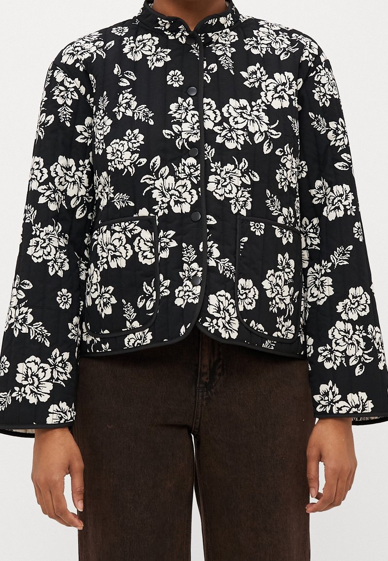 Veste matelassée noire avec motif floral blanc, boutons-pression sur le devant, deux grandes poches avant, portée avec un pantalon marron foncé.