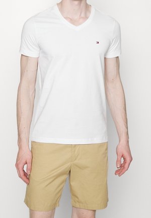 Mann trägt weißes V-Ausschnitt-T-Shirt mit kleinem Logo auf der Brust und beige Shorts vor einfarbigem Hintergrund.