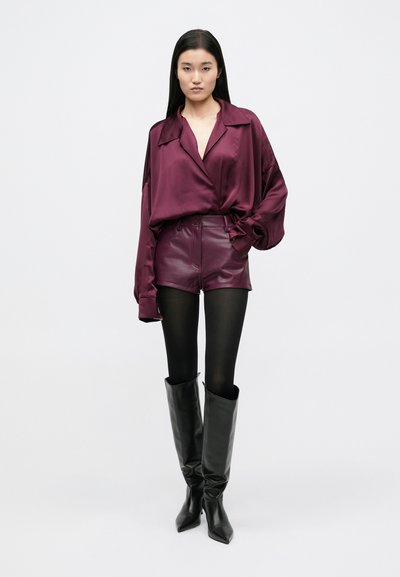 Vinjen satenasta bluza z širokim ovratnikom, kombinirana z usnjenimi kratkimi hlačami z visokim pasom, črnimi nogavicami in sijočimi črnimi škornji do kolen.