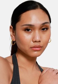 Goldene Creolen mit glatter Oberfläche, runder Form und leichter Dicke. Das Model hat einen natürlichen Make-up-Look und trägt ein dunkles Halsteroberteil.