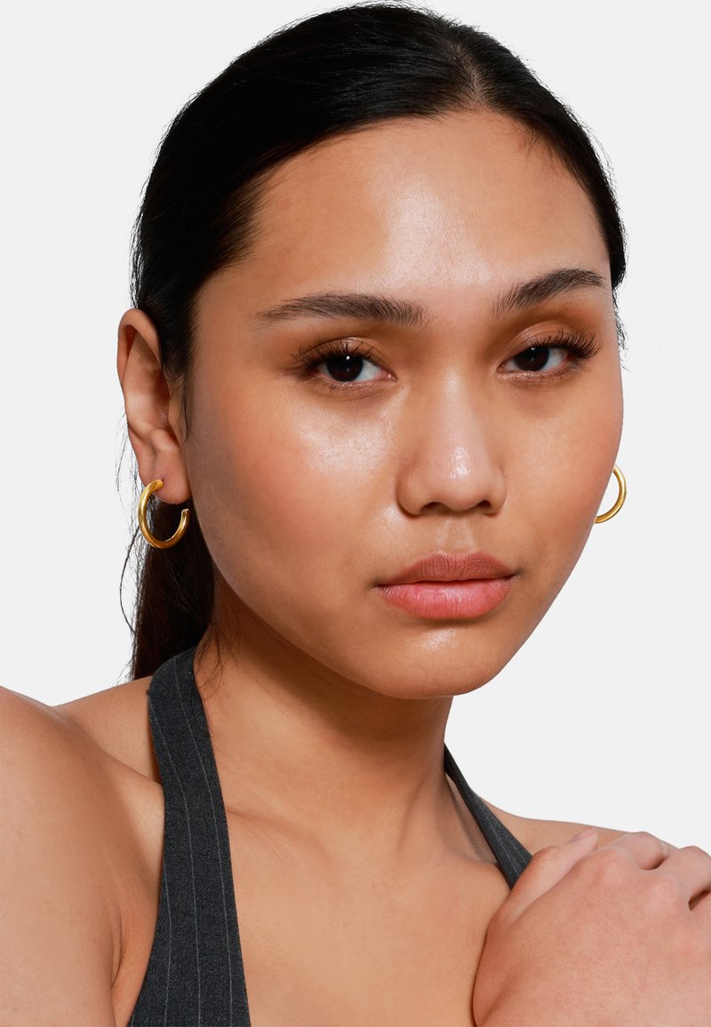 Goldene Creolen mit glatter Oberfläche, runder Form und leichter Dicke. Das Model hat einen natürlichen Make-up-Look und trägt ein dunkles Halsteroberteil.