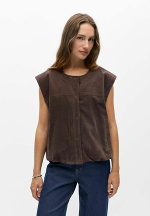 Kvinde med langt hår iført en ærmeløs brun kordfløjls-top og blå jeans, der står mod en ensfarvet hvid baggrund.