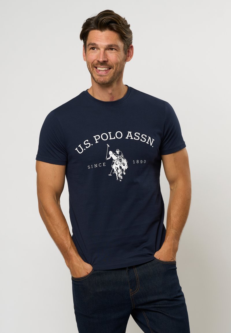 Mørkeblå kortermet t-skjorte laget av bomull, med en hvit grafisk logo som viser en polo-scene og teksten "U.S. Polo Assn. Siden 1890."