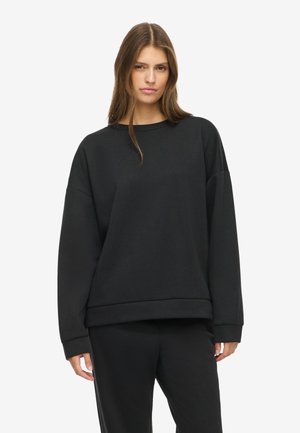 Femme aux longs cheveux bruns portant un sweat-shirt noir ample et un pantalon noir assorti, debout devant un fond blanc uni.