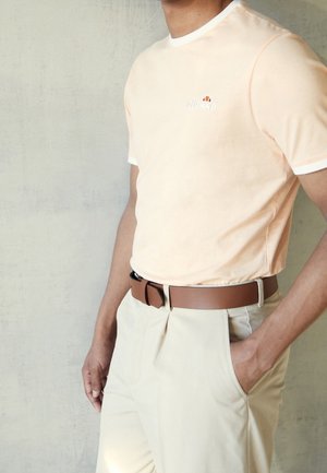 Mężczyzna w jasnopomarańczowej koszulce z krótkim rękawem z białymi obszyciami, beżowych spodniach i brązowym pasku, stojący z jedną ręką w kieszeni.