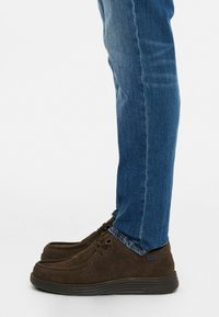 Zapatos de ante marrón con diseño de cordones y suela de goma, combinados con jeans de mezclilla azules, que muestran detalles en los tobillos y costuras.