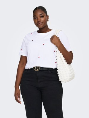 Kvinde iført hvid T-shirt med små røde hjerter, sorte bukser, guldringearrøreringe, sort bælte og bærende en hvid skuldertaske med struktur.