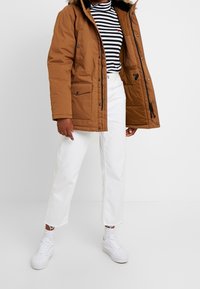 Braune Steppjacke mit Kunstpelzbesatz, zwei Fronttaschen, getragen über einem schwarz-weißen gestreiften Hemd, kombiniert mit weißen Hosen und Sneakers.