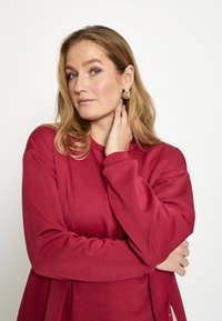 Pehmeast kangast punane pika varrukaga pluus, lõdva lõikega ja külgnevõludega. Aksessuaarideks hõbedased kõrvakad, kandmise poos on vaba ja lihtne.