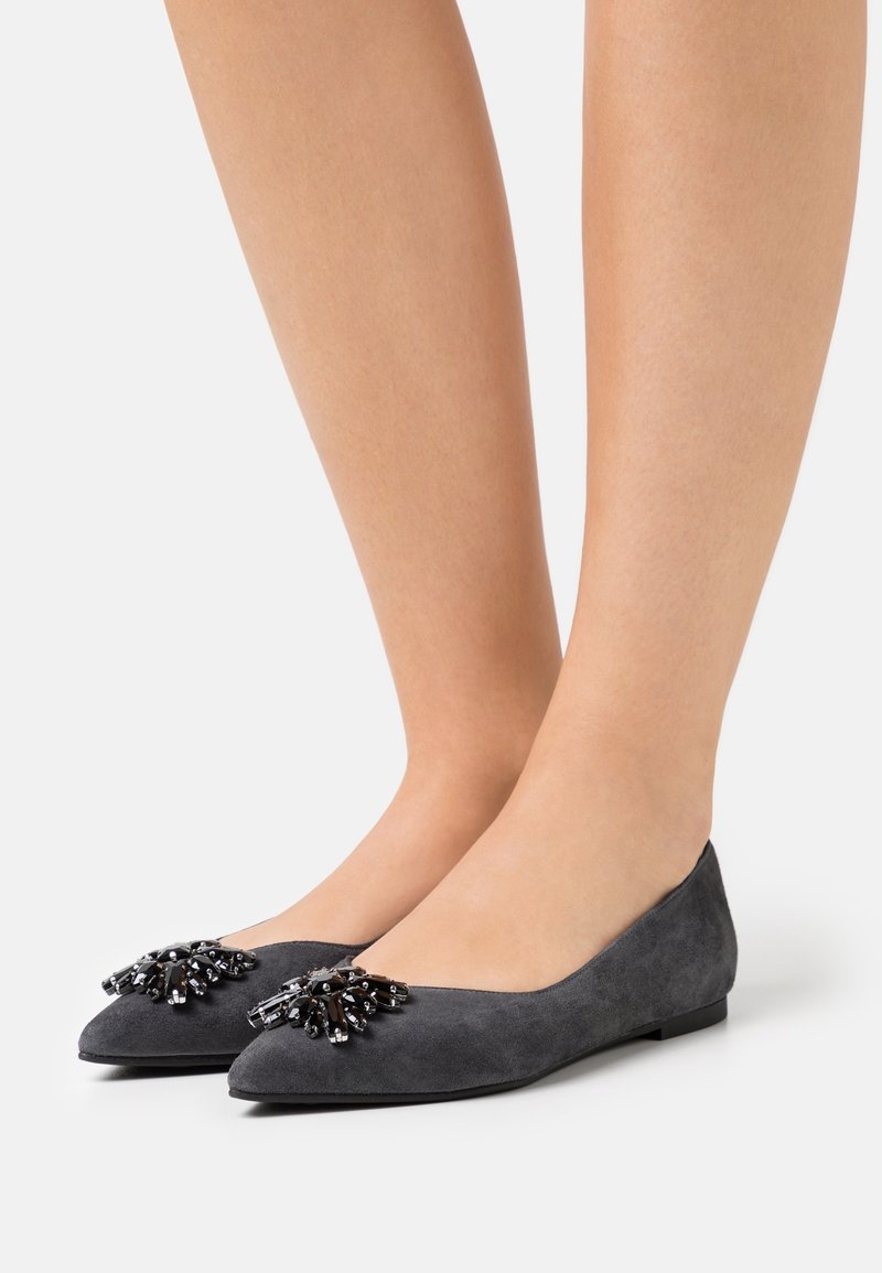 Ballerines en daim gris à bout pointu avec des ornements de perles noires et argentées, portées pieds nus sur fond blanc.