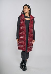 Burgundowa pikowana kamizelka z błyszczącym wykończeniem, do kolan, z zamkiem błyskawicznym i kapturem. W połączeniu z czarną sukienką z długim rękawem i masywnymi czarnymi butami.