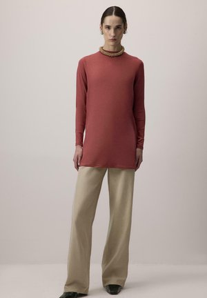 Γυναίκα στέκεται μπροστά σε απλό φόντο φορώντας μακρυμάνικη τουνίκ σε χρώμα σκουριάς, μπεζ παντελόνι με φαρδιά μπατζάκια, μαύρα παπούτσια και χοντρό κολιέ με χάντρες.