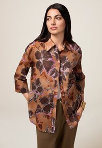 Cămașă maro cu flori, cu manșete de trei sferturi, având modele violet și negre, un guler ascuțit și o croială lejeră.