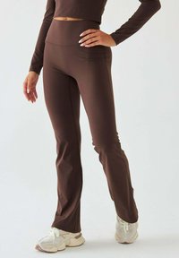 Brązowe spodnie dzwony wykonane z elastycznego materiału z wysokim stanem. Gładka tekstura i dopasowana sylwetka, zestawione z białymi butami sportowymi.