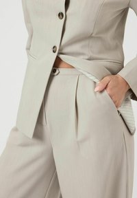 Ljusbeige kostym med en skräddarsydd kavaj med en knapp och vida byxor med veck; randigt tyg synligt från fickan.
