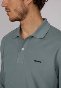 Polo en teal atténué, fabriqué en tissu texturé. Il dispose d'une patte de boutonnage à trois boutons et d'un petit motif d'ours brodé en marine sur la poitrine.