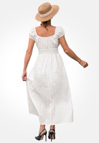 Vestido maxi blanco con textura, mangas cortas abullonadas y cinturilla fruncida. Combinado con zapatos de tacón negros y un sombrero de paja color marrón.