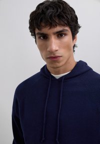 Young man with dark curly hair wearing a navy blue hoodie over a white shirt, looking directly at the camera against a plain light background.