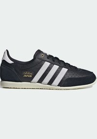 Black sneaker with white stripes, made of leather and suede. Features a perforated design, gold logo detail, and a textured rubber outsole.