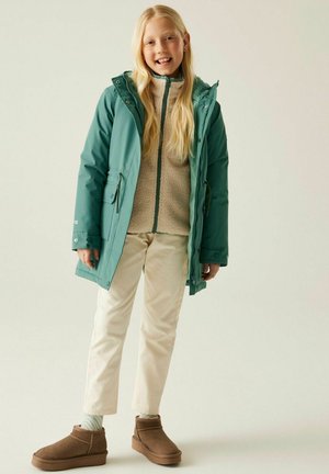Młoda dziewczyna o długich blond włosach, ubrana w turkusową kurtkę i beżowe spodnie, uśmiechnięta, stojąca na tle jednolitego, jasnego tła.