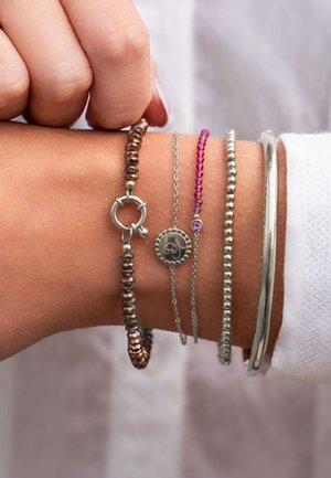 Nadgarstek z pięcioma bransoletkami: brązowa z koralików, srebrny łańcuszek z okrągłym zawieszkiem, różowa z koralików, srebrna z koralików oraz gładka srebrna bransoletka.