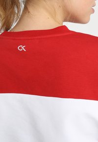 T-shirt in cotone rosso e bianco con scollatura rotonda e un piccolo logo bianco sul retro, texture liscia e design sportivo.
