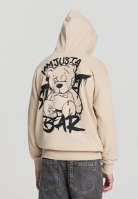 Felpa beige con una grafica nera di un orso dei cartoni animati sulla schiena, che presenta il testo "SONO SOLO UN ORSO." Tessuto morbido, design casual.