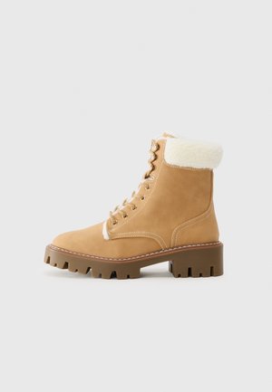 Bottines en suède beige avec un col en fausse fourrure blanche, bout rond, solide talon bloc et œillets en métal pour les lacets. Semelle brune striée.