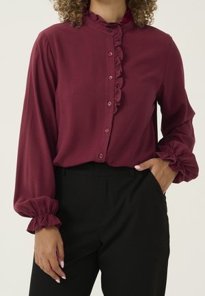 Vrouw draagt een bordeauxrode blouse met lange mouwen en ruches op de knoopsluiting en manchetten, gecombineerd met zwarte broek met hoge taille, hand in de zak.