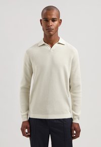 Lyst beige strikket langermet poloshirt med V-hals og kontrastfarge på kragen. Har et strukturert mønster og ribbestrikkede mansjetter.