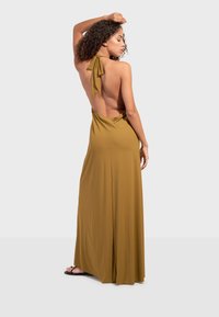 Femme aux cheveux bouclés posant avec un bras levé, portant une robe longue dos nu à col halter de couleur vert olive et des sandales noires.