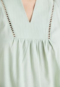 Light green blouse featuring a V-neck, subtle stripes, and cut-out detailing along the neckline. Soft texture with gathered fabric below the chest.