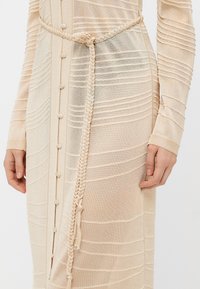 Beige knitted dress featuring textured horizontal stripes, front buttons, and a braided tie at the waist. Long sleeves complete the design.
