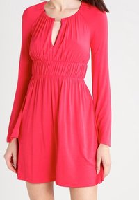 Robe rose à manches longues avec un décolleté en forme de clé et une taille froncée. Fabriquée dans un tissu lisse et léger, elle possède une jupe évasée.