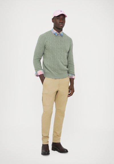 Camisola verde de malha com padrão trançado, gola de camisa aos quadrados, calças cargo bege e botins pretos até ao tornozelo. Boné rosa com acento de logo. Fundo minimalista.