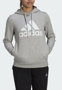Šedá mikina s kapucí vyrobená z měkkého materiálu, s velkým bílým logem Adidas na přední straně, klokaní kapsou a nastavitelnou kapucí na šňůrky.