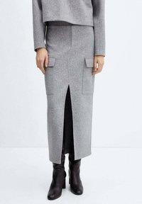 Jupe en mélange de laine grise avec une fente à l'avant, deux poches avant et une coupe ajustée. Associée à des bottes noires à talons mi-mollet.