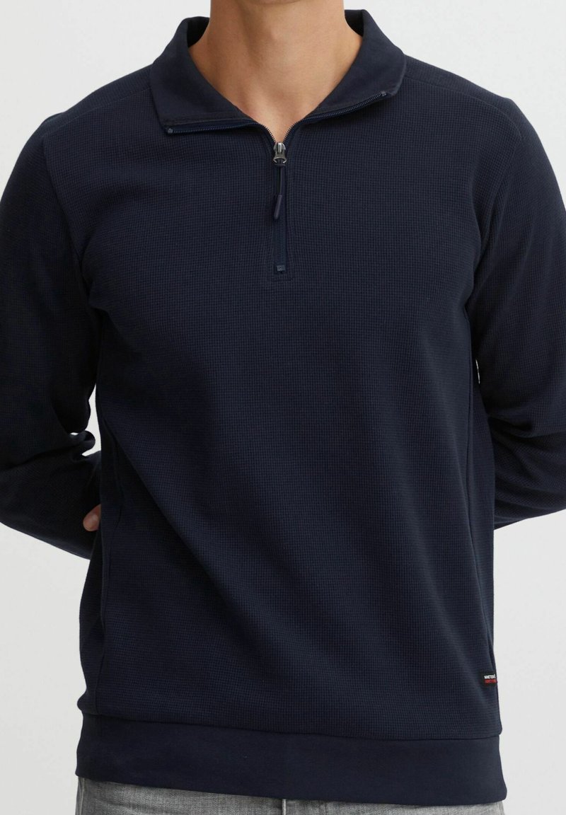 Pull en navy sombre en tissu texturé avec un col zippé quart et un ourlet côtelé, doté de manches longues et d'un petit logo de marque.