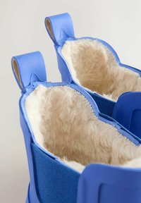 Mėlyni batai su minkštu, kreminės spalvos vilnos pamušalu ir guminėmis išorinėmis dalimis. Yra dvi viršutinės rankenos patogiam dėvėjimui. Tekstūruotos audinio detalės.