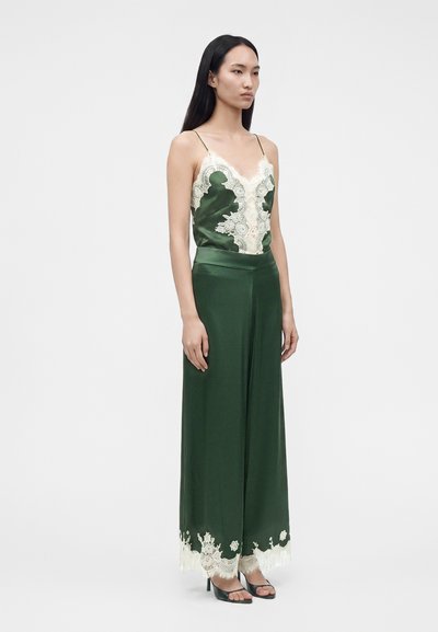 Femme portant un caraco en satin vert foncé et un pantalon large avec des détails en dentelle blanche, debout bras détendus devant un fond uni.