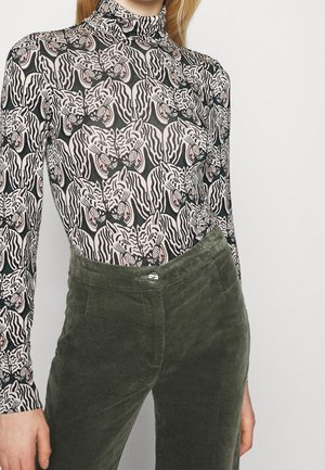 Frau mit hochgeschlossenem Oberteil mit schwarzem und weißem Tigerkopfmuster und olivgrünen Samthosen.