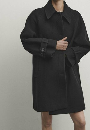 Personne portant un manteau noir surdimensionné avec de larges manches et des poignets boutonnés, debout devant un fond clair uni.