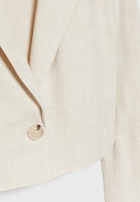 Blazer court beige clair en tissu texturé. Il présente des revers crantés et une fermeture à un seul bouton, avec un motif tissé subtil.