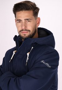 Giacca a vento blu navy con cappuccio e zip, tessuto texturizzato, cordini bianchi con dettagli in legno e logo sul lato sinistro della manica.