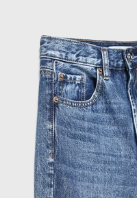 Lähetetyn denim-farkun lähekuva, jossa näkyy etuvasen tasku kolikoille, vyölenkki, metalliset niitit ja nappi vaaleanharmaalla taustalla.
