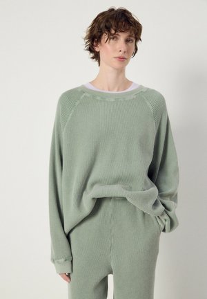 Persona con capelli castani corti che indossa un maglione ampio verde chiaro a trama a nido d'ape e pantaloni abbinati su uno sfondo chiaro e semplice.