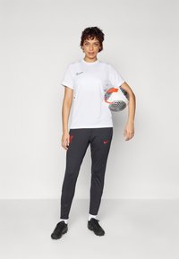 Balta Nike sportinė marškinė su juodu logotipu, derinama su tamsiai pilkomis Nike kelnėmis su raudonu akcentu. Žaidėjas laiko raštuotą futbolo kamuolį.