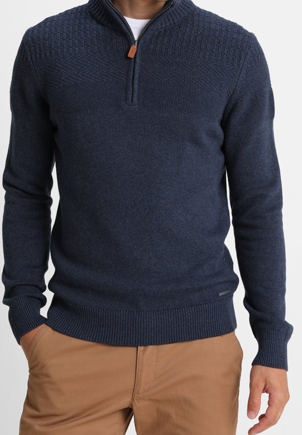 Homme portant un pull en maille texturée bleu marine avec fermeture éclair quart, sur une chemise blanche et un pantalon marron clair, les mains dans les poches.