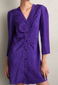 Lila klänning med knapplist, V-ringning, trekvartslånga ärmar, textil med struktur som har ett mönster av cirklar över hela ytan och rynkdetaljer.